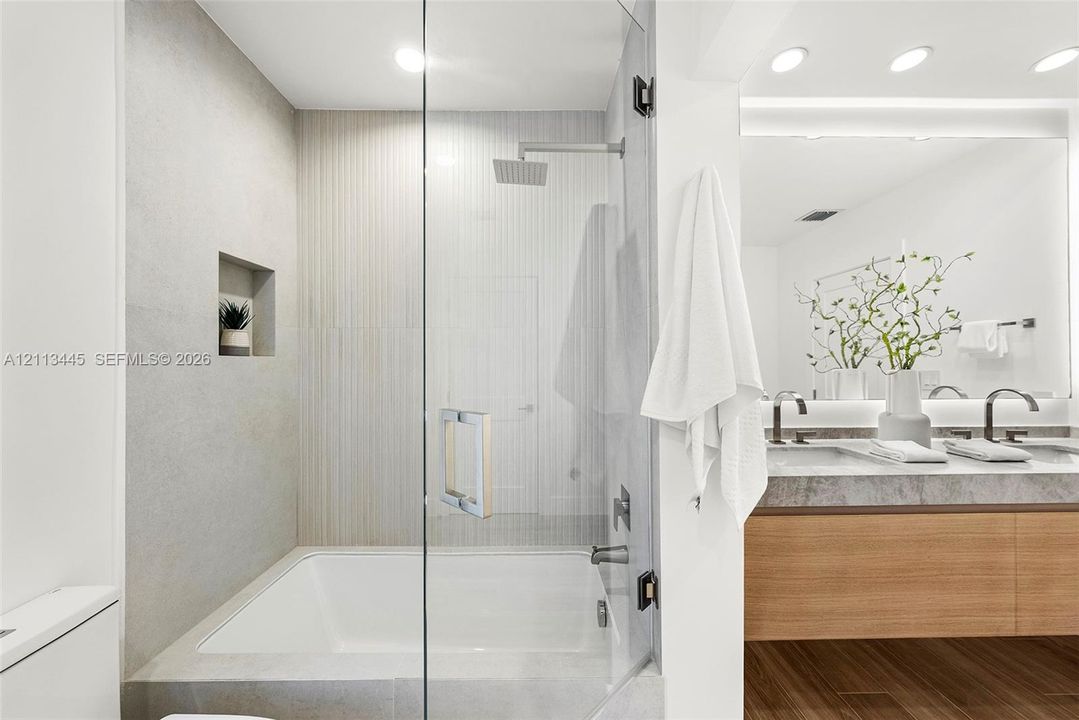 Shower and tub combination featuring glass enclosure, rain-style shower head, built-in recessed wall niche, dual-sink vanity with stone-finish countertop, and wood-finish flooring
