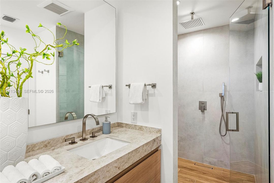 Bathroom featuring a stone-surface vanity with an integrated sink, wood-finish cabinetry, a large wall-mounted mirror, and a walk-in glass-enclosed shower with a rainfall showerhead and wall-mounted hand sprayer