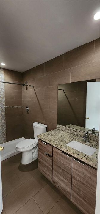 Bathroom featuring a wood-finish vanity with a stone countertop and rectangular undermount sink, ceramic tile flooring, and floor-to-ceiling tile shower surround