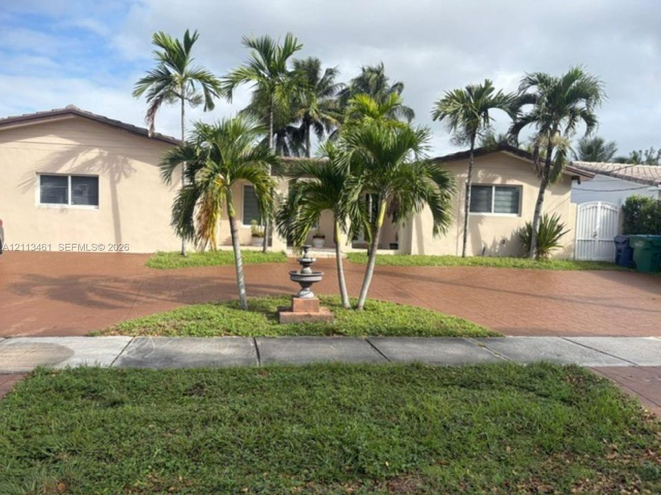 Single-story residence featuring a paver driveway, mature palm trees, a decorative central stone fountain, and a white arched side gate