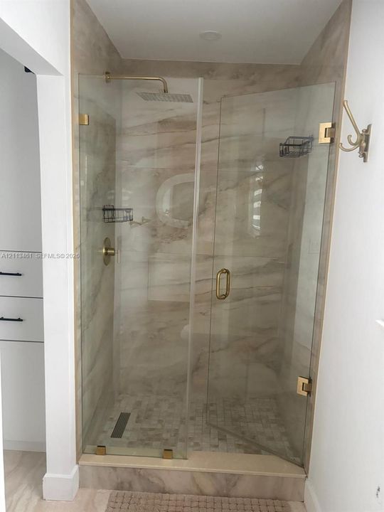 Walk-in shower featuring large-format stone-pattern wall tile, mosaic stone floor tile, frameless glass enclosure, brass-finish rainfall shower head, and matching brass-finish hardware