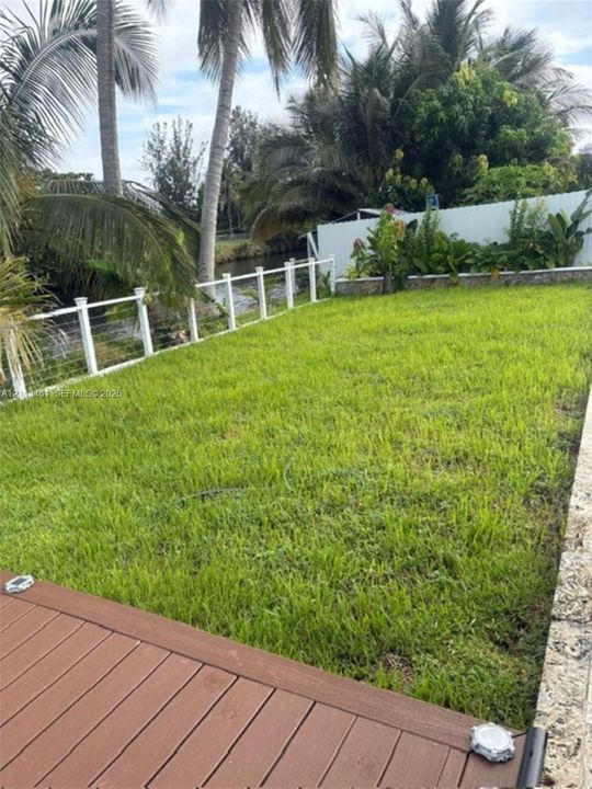 Backyard featuring a wood-finish deck, natural grass lawn, white post-and-cable perimeter fencing, stone retaining wall, and mature palm trees