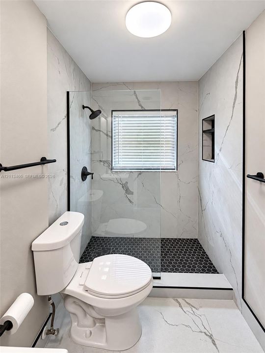 Full-height stone-finish wall tile, frameless glass shower enclosure with matte black hardware, black hexagonal floor tile, recessed shower niche, and modern flush-mount ceiling lighting