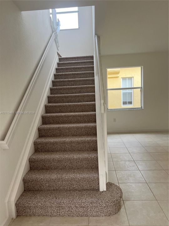Staircase featuring wall-to-wall carpeting and white wooden handrails, complemented by ceramic tile flooring and windows providing natural illumination