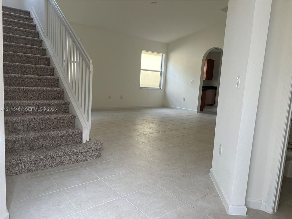 Open interior featuring neutral ceramic tile flooring, a carpeted staircase with white balusters and handrail, and an arched doorway
