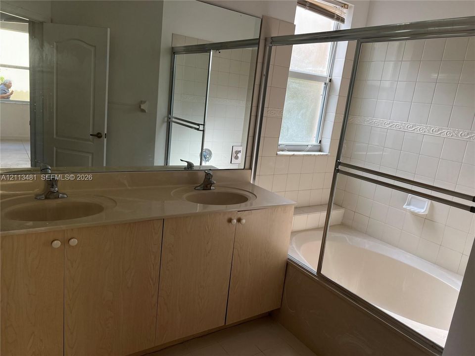 Bathroom featuring a dual-sink vanity with a solid-surface countertop, full-width wall mirror, soaking tub with a tiled surround, and a sliding glass shower enclosure