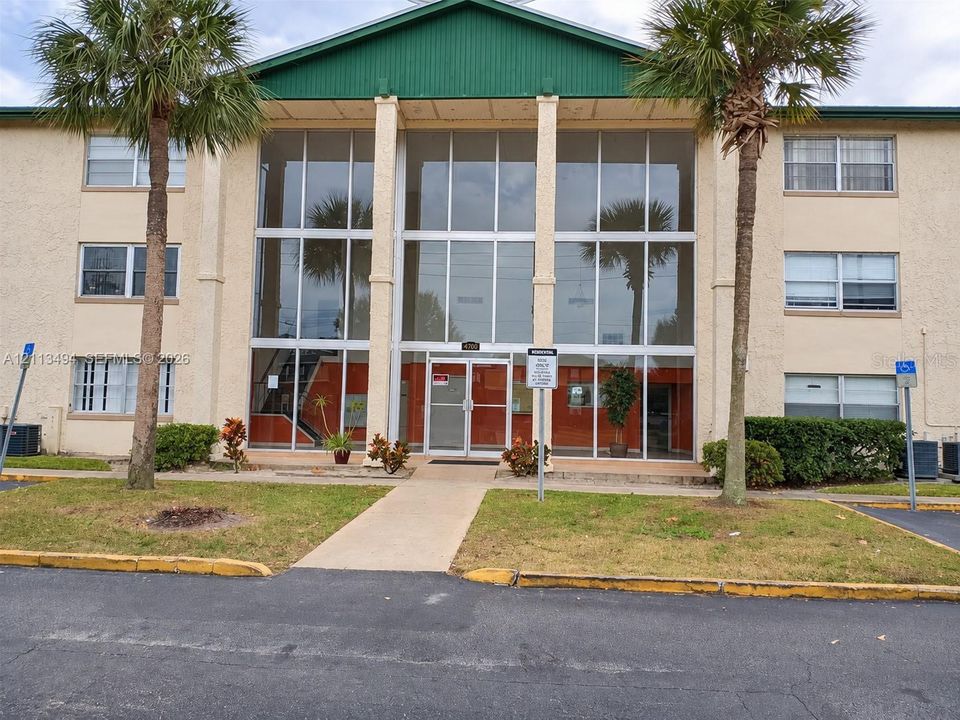 Multi-story exterior featuring a stucco facade, expansive floor-to-ceiling glass windows, a prominent green metal roof gable, and mature palm landscaping