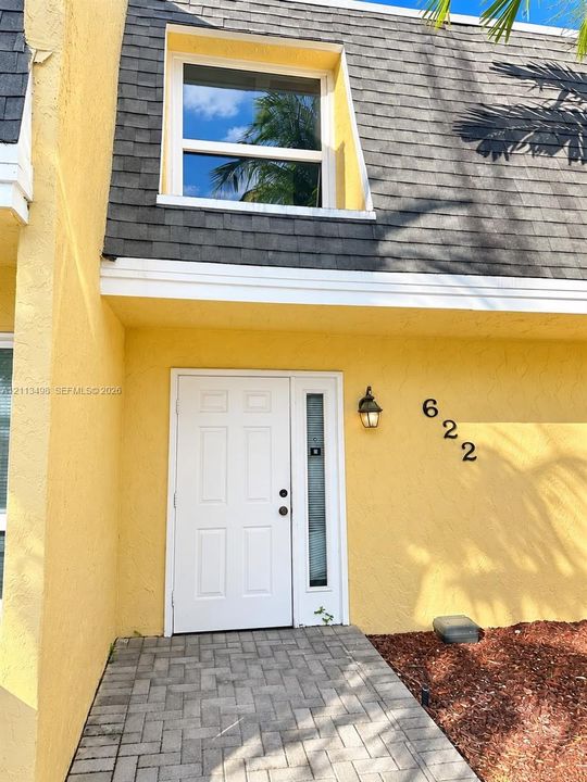 Exterior entrance featuring a paver walkway, white paneled door with glass sidelight, dark shingle mansard roof, and wall-mounted exterior lighting