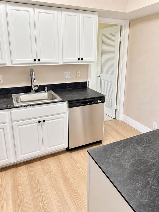 Kitchen featuring white Shaker-style cabinetry, dark stone-finish countertops, an undermount stainless steel sink with a high-arch faucet, a stainless steel dishwasher, and wood-finish flooring