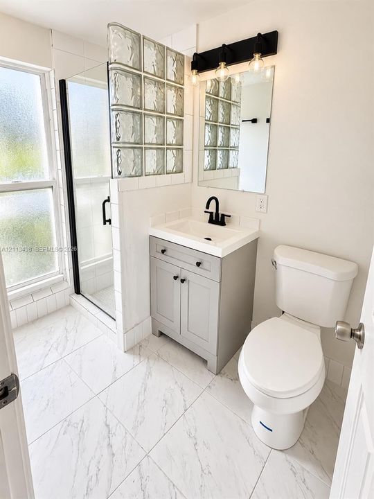 Bathroom featuring a walk-in shower with a glass block partition, stone-finish tile flooring, a gray vanity with a rectangular basin, and a matte black faucet