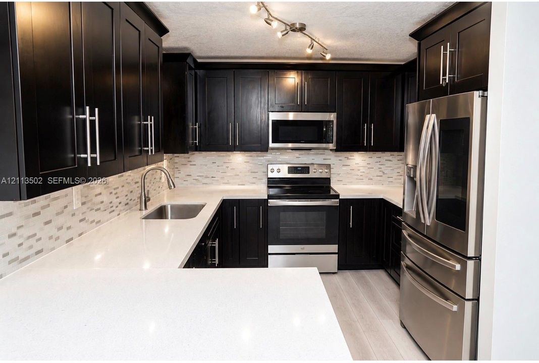 Kitchen featuring dark shaker-style cabinetry, white quartz countertops, multi-toned linear tile backsplash, stainless steel appliances, and track lighting