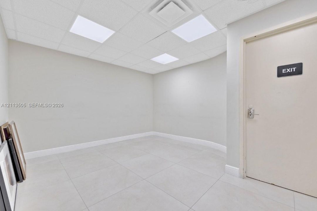 Neutral-toned interior space featuring oversized ceramic tile flooring, recessed drop-ceiling lighting panels, and a solid-core door with metallic hardware