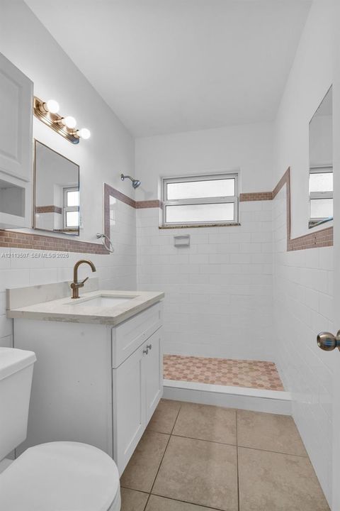 Bathroom featuring a white vanity with a neutral stone countertop, bronze-finish faucet, white subway tile walls with decorative border accents, walk-in shower with mosaic tile flooring, and large-format tile flooring
