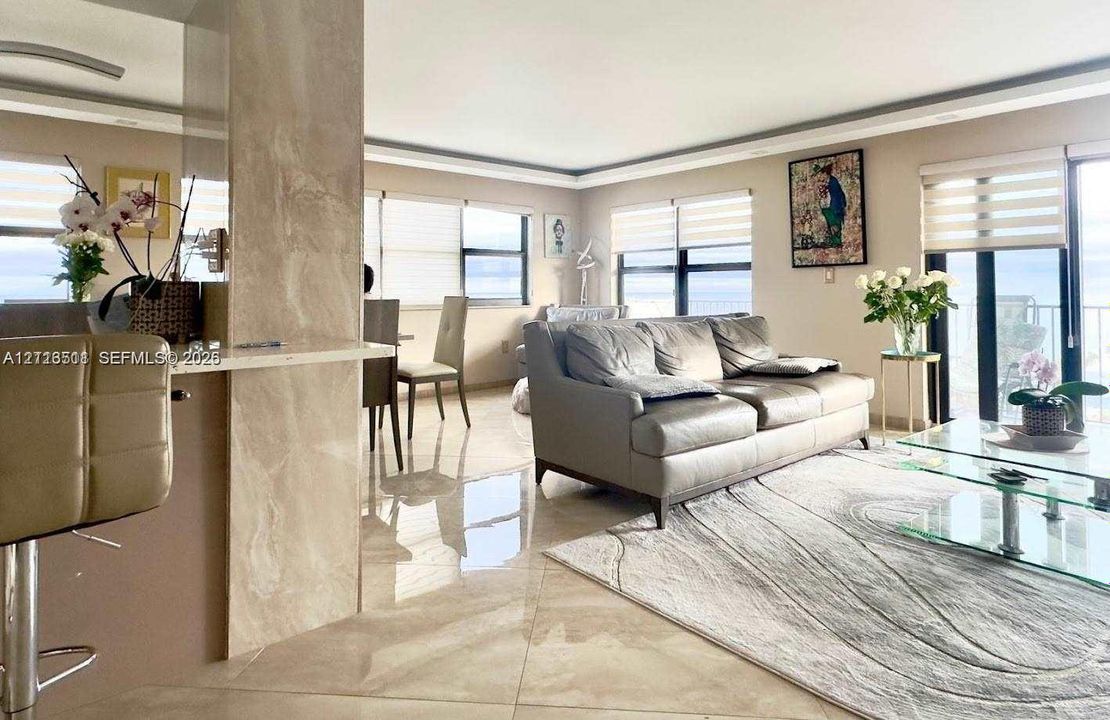 Open-concept living space featuring large-format polished tile flooring, natural stone-faced breakfast bar, recessed tray ceiling, and expansive windows with custom roller shades