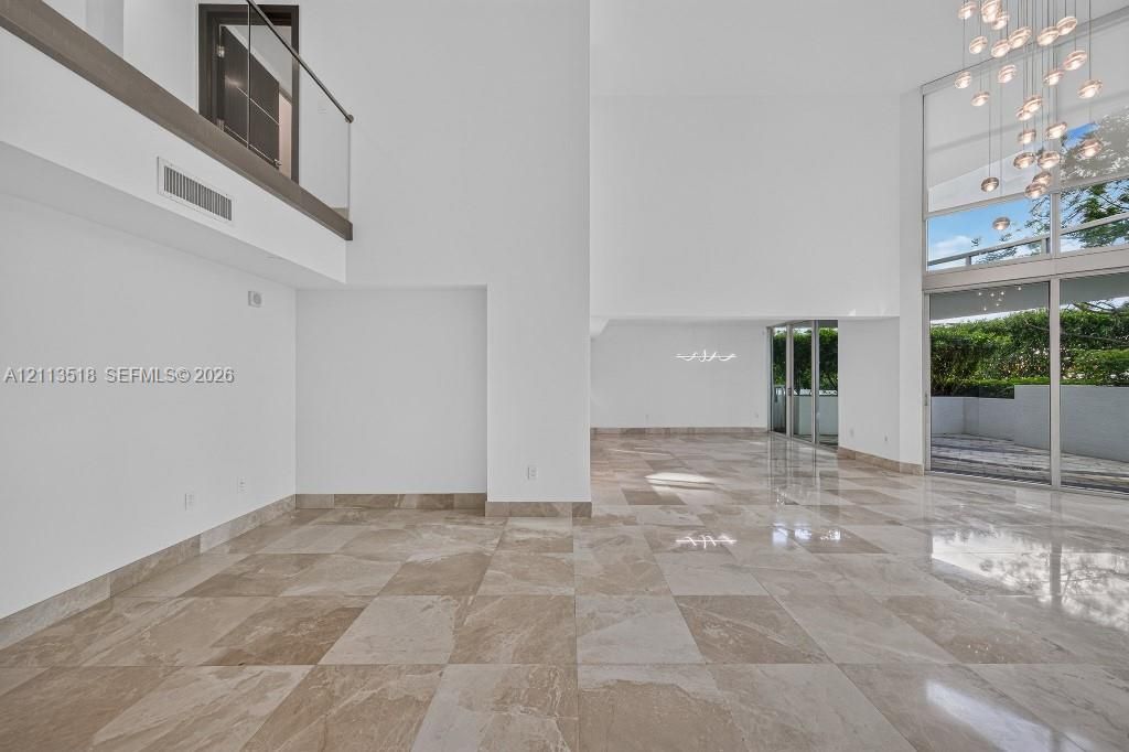 Double-height ceilings featuring floor-to-ceiling windows, polished stone flooring, a multi-pendant chandelier, and a glass-paneled mezzanine railing