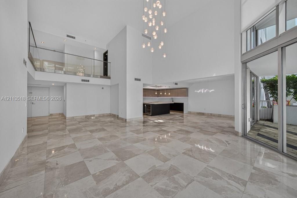 Open-concept interior featuring double-height ceilings, stone-finish tile flooring, a glass-panel mezzanine railing, multi-light pendant chandelier, and expansive floor-to-ceiling sliding glass doors