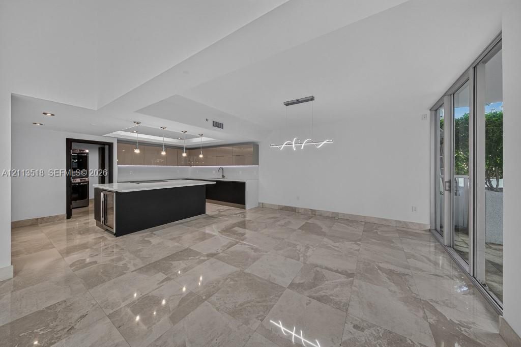 Open-concept area featuring oversized neutral-toned tile flooring, sleek flat-panel cabinetry, a central kitchen island with a stone-finish countertop, minimalist pendant lighting, and floor-to-ceiling glass sliding doors