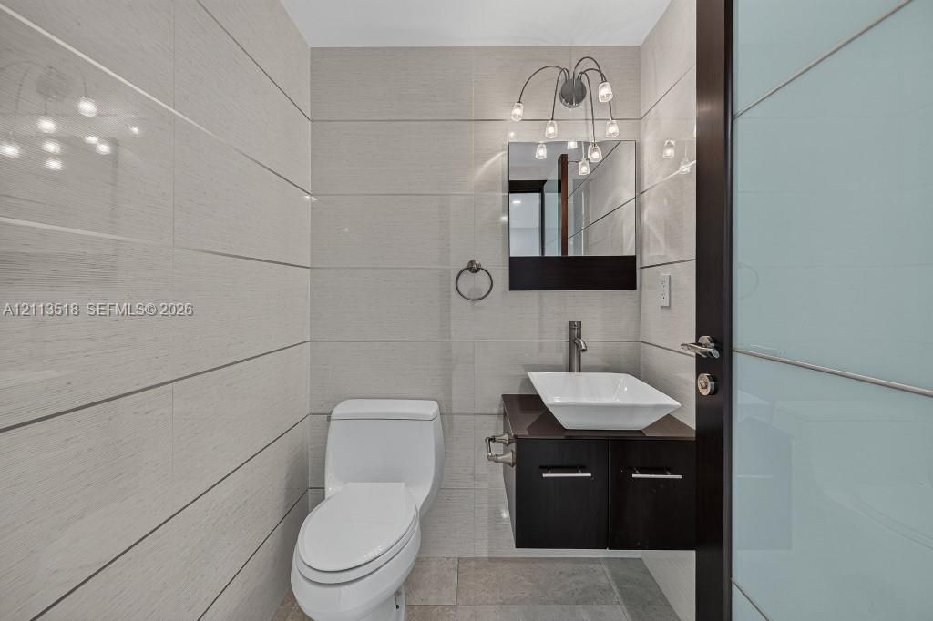 Bathroom featuring floor-to-ceiling large-format tile, wall-mounted dark wood vanity with a white vessel sink, multi-bulb wall-mounted lighting, and a frosted glass panel door