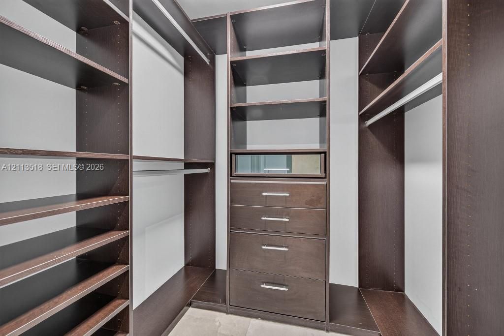 Custom walk-in storage system featuring dark wood-finish cabinetry, built-in shelving, integrated hanging rods, and a centralized unit with four drawers and a glass-front display compartment