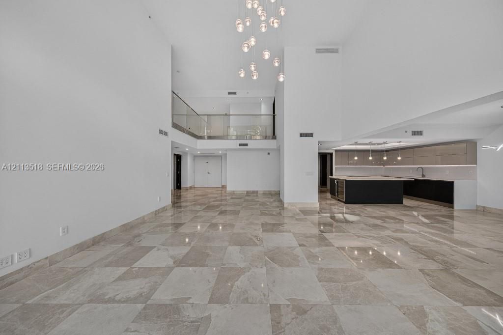 Expansive interior featuring high ceilings, stone flooring, a multi-tiered glass pendant chandelier, a glass-railed mezzanine, and kitchen cabinetry with integrated lighting