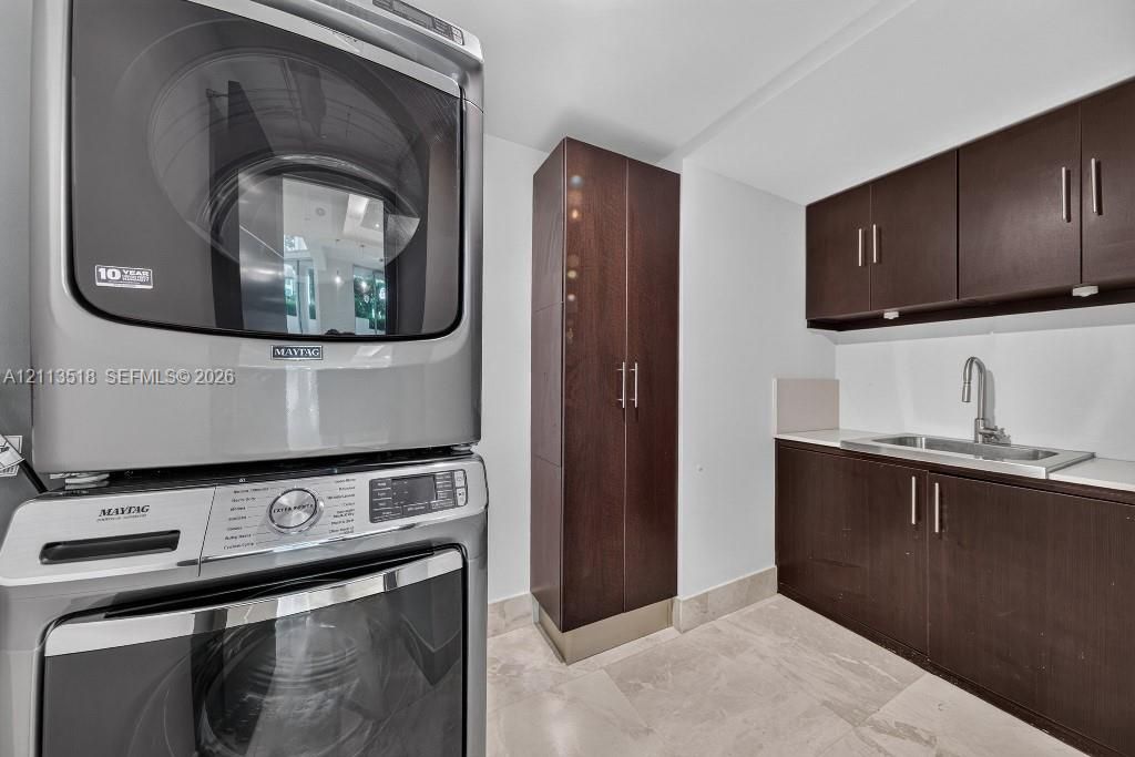 Stacked high-efficiency metallic laundry center, custom dark wood-finish cabinetry, floor-to-ceiling storage pantry, stainless steel undermount sink with a gooseneck faucet, and stone-finish flooring