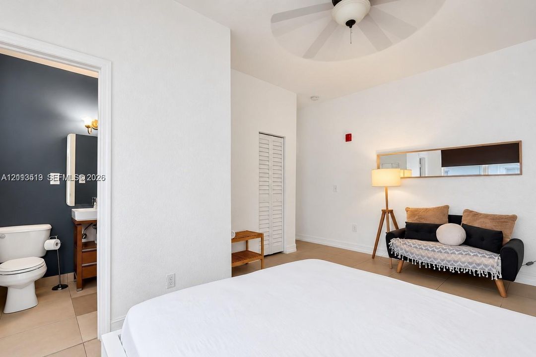 Room featuring high ceilings with a mounted ceiling fan, neutral wall finishes, tile flooring, a louvered-door closet, and an attached bathroom with a dark accent wall and a built-in vanity