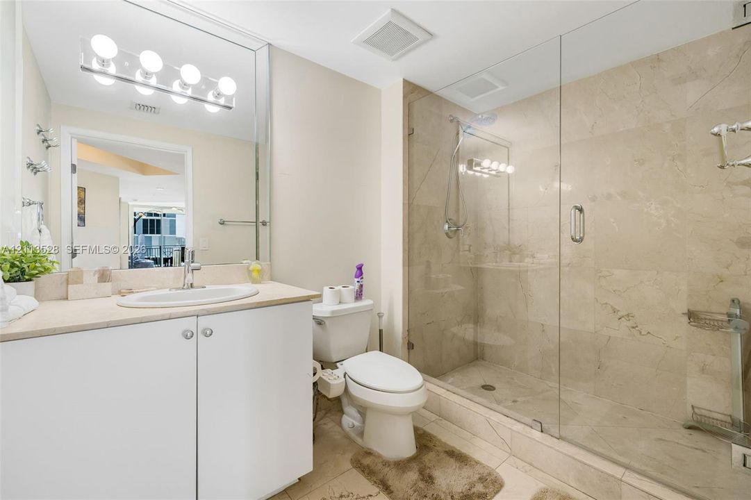 Bathroom featuring a white vanity with an integrated sink, a full-width vanity mirror, a walk-in shower with a glass enclosure and stone-finish wall tile, and polished chrome hardware