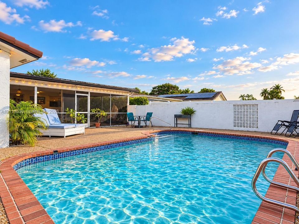 In-ground swimming pool with brick coping, surrounded by a textured aggregate patio and enclosed by a masonry privacy wall featuring integrated glass block accents, complemented by a screened-in lanai