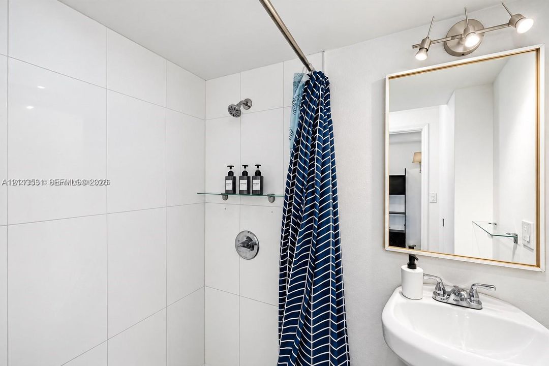 Wall-mounted glass shelf, white tile shower surround, polished chrome plumbing fixtures, rectangular mirror with metallic frame, and wall-mounted spotlight vanity lighting