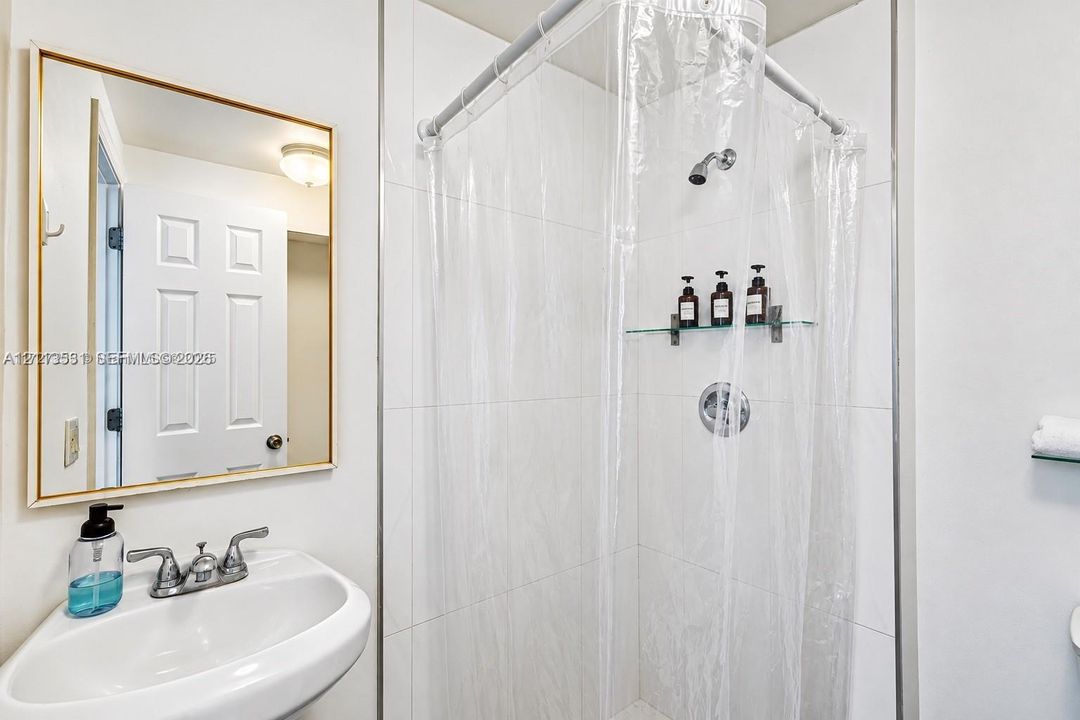 Wall-mounted basin sink with a dual-handle chrome faucet, rectangular gold-framed mirror, and floor-to-ceiling tiled shower enclosure featuring a glass shelf