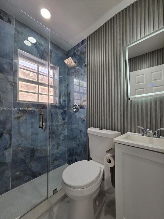 Glass-enclosed shower with blue-toned large-format wall tile, ribbed wall paneling, vanity with integrated basin, and built-in illuminated vanity mirror