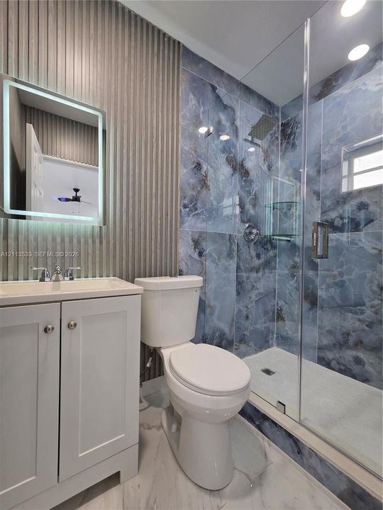 Bathroom featuring a glass-enclosed shower with blue veined tile surround, vertical slatted accent wall, integrated LED-lit vanity mirror, and white shaker-style cabinetry
