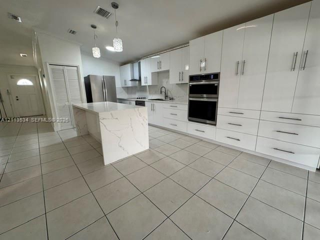 Kitchen featuring white shaker-style cabinetry, an expansive center island with stone-patterned surfaces, a double wall oven, stainless steel refrigerator, and large-format tile flooring