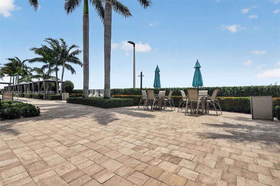 Expansive paver patio featuring manicured hedges, mature palm trees, and structural cabanas