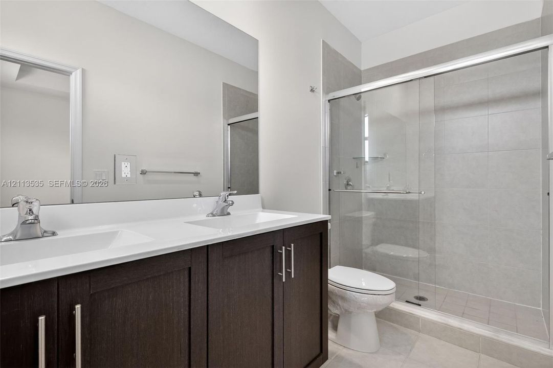 Dark wood-finish double vanity with white countertop, dual undermount sinks, large wall-mounted mirror, and glass-enclosed shower with tiled surround