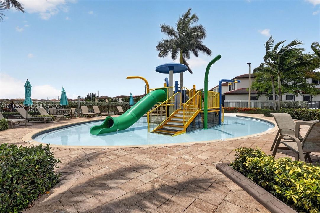 Splash pad featuring a central play structure with a slide, water features, concrete paver decking, and mature palm landscaping