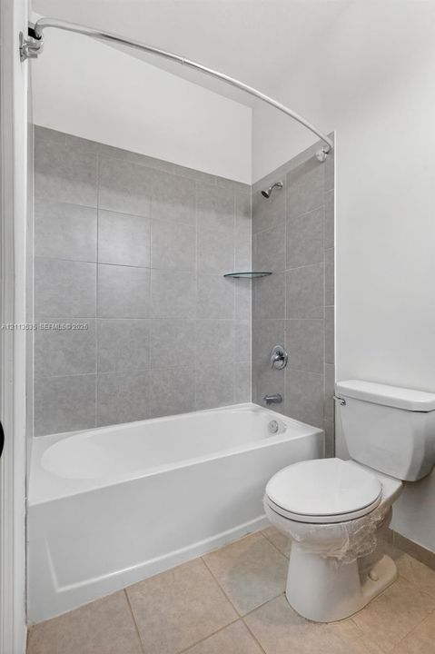 Full bathtub with shower combination, gray tile surround, integrated corner glass shelf, and standard white toilet