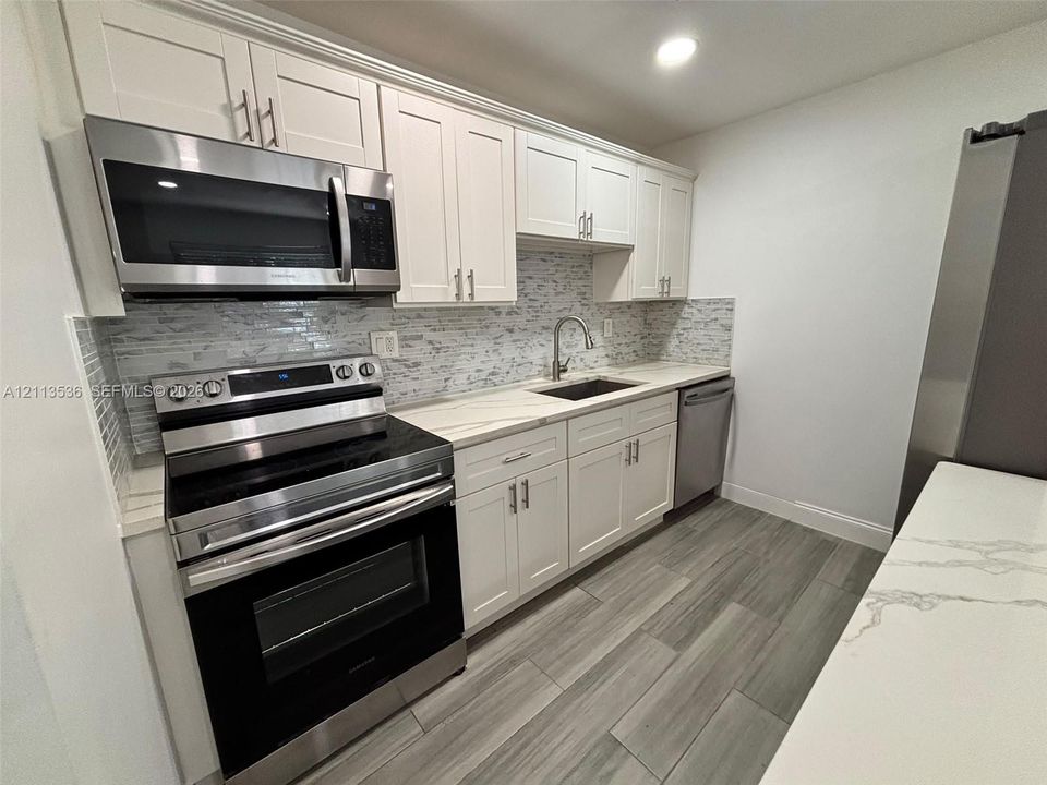 Kitchen featuring white Shaker-style cabinetry, stainless steel appliances, quartz countertops, a stone-tile backsplash, and wood-finish flooring