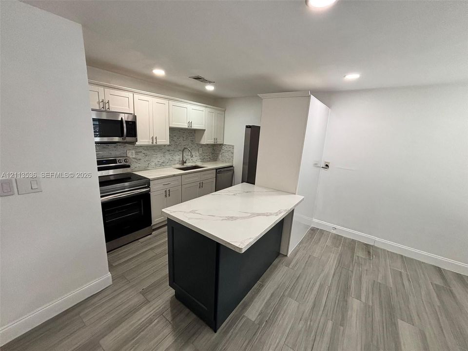 Kitchen featuring white cabinetry, an island with a stone-composite countertop, stainless steel appliances, a tile backsplash, and wood-finish flooring