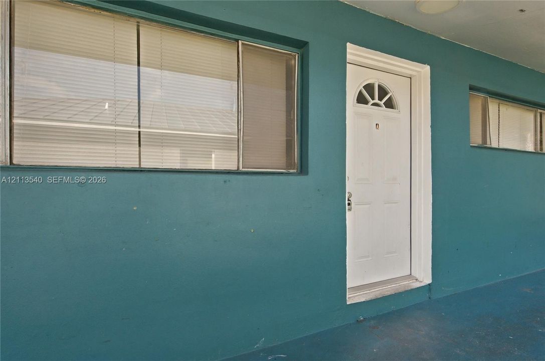 Exterior entryway featuring teal stucco siding, a white six-panel door with a semi-circle transom window, and multi-pane windows with horizontal blinds