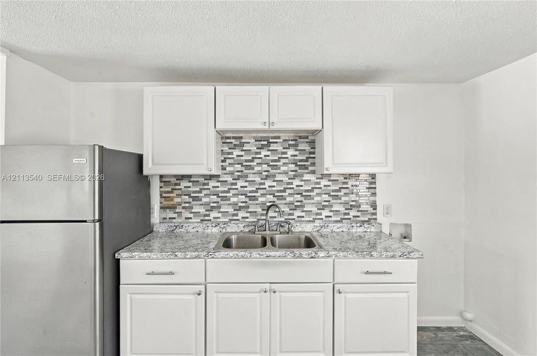 Kitchen area featuring white shaker-style cabinetry, stone-patterned countertops, a multi-tonal mosaic tile backsplash, a stainless steel dual-basin sink with a gooseneck faucet, and a stainless steel refrigerator