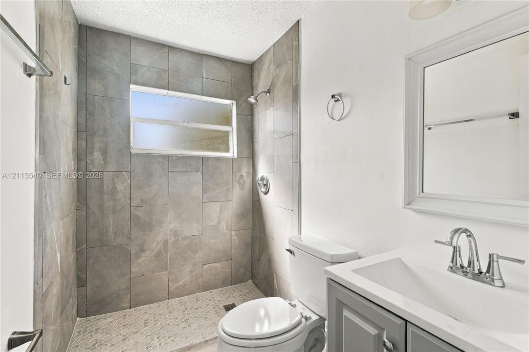Bathroom featuring a walk-in shower with large-format stone-finish tile surround, pebble-texture flooring, a wall-mounted rectangular window, vanity with integrated basin, and polished chrome fixtures