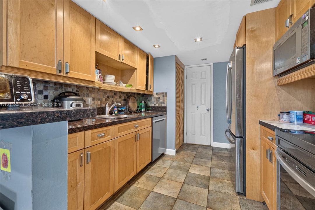 Kitchen featuring natural wood cabinetry, mosaic tile backsplash, stone-finish countertops, stainless steel appliances, and tiled flooring