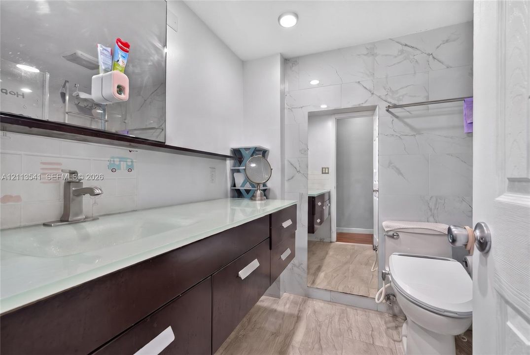 Bathroom featuring an extended dark-finish vanity with an integrated glass countertop, large-format veined wall tile, wood-finish flooring, and a sleek faucet fixture
