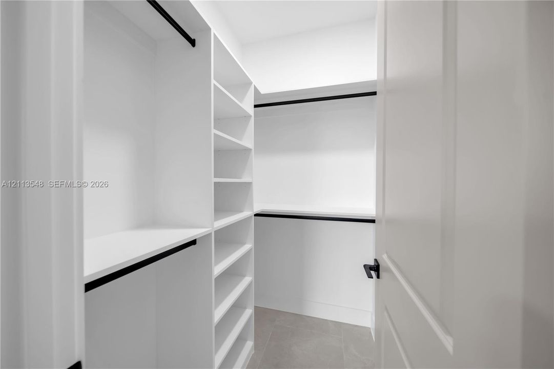 Walk-in closet featuring white built-in shelving, matte black hanging rods, tile flooring, and a solid-core door with matte black hardware