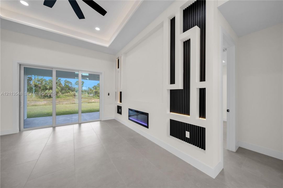 Tray ceiling with recessed lighting, large-format tile flooring, wall-mounted electric fireplace, custom geometric wall accent with vertical slat detailing, and multi-panel sliding glass doors