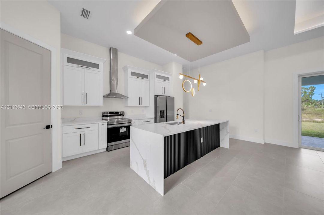 Kitchen featuring an oversized island with a waterfall-edge countertop, black slatted cabinetry, stainless steel appliances, recessed ceiling detail, and large-format tile flooring