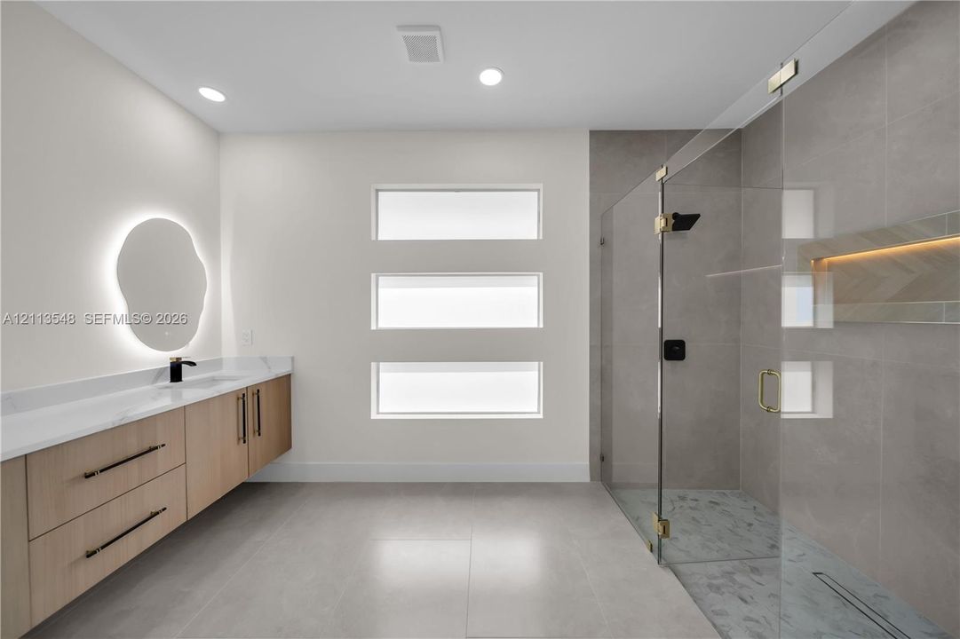 Floating wood-finish vanity with stone countertop, frameless glass walk-in shower with recessed tiled niche, and triple-stacked horizontal windows