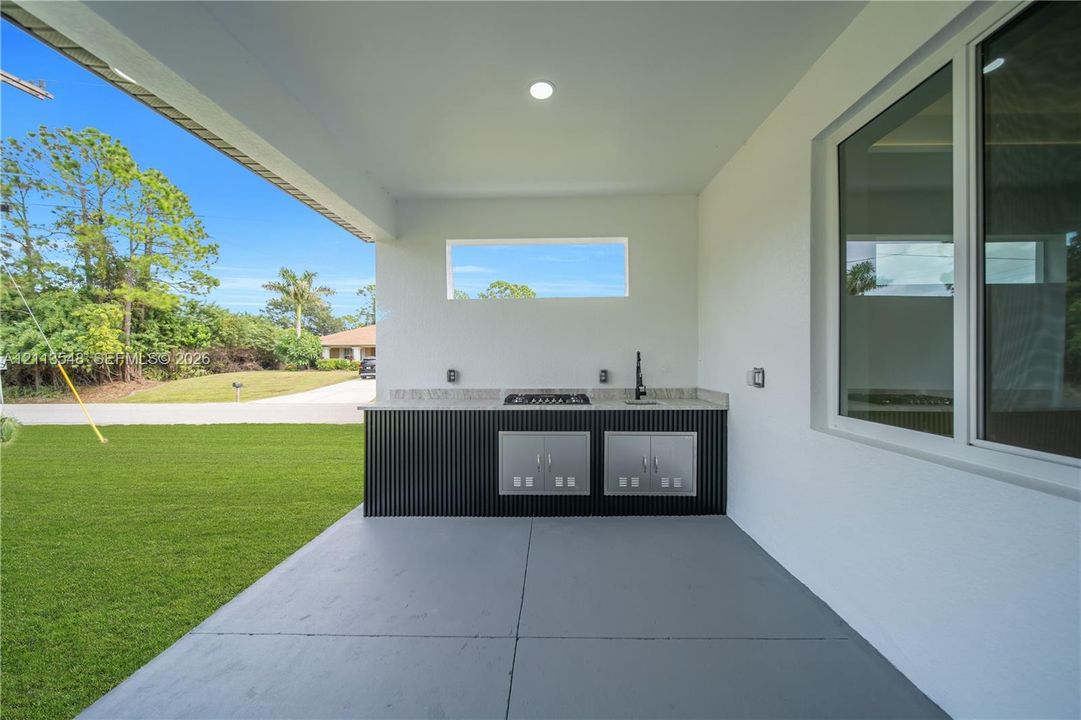 Covered outdoor kitchen featuring stainless steel built-in cabinetry, a stone-surfaced countertop, a cooktop, a sink with a gooseneck faucet, and concrete flooring
