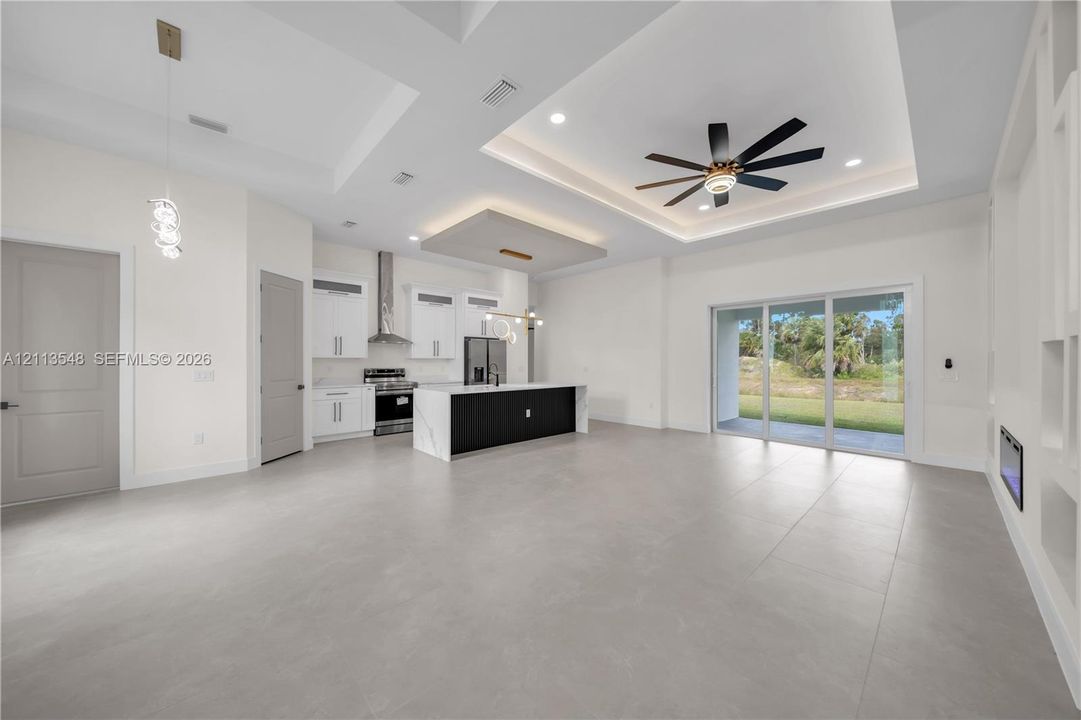 Open-concept floor plan featuring large-format tile flooring, tray ceilings with recessed lighting, a center island with contrasting cabinetry, a built-in linear fireplace, and multi-panel sliding glass doors